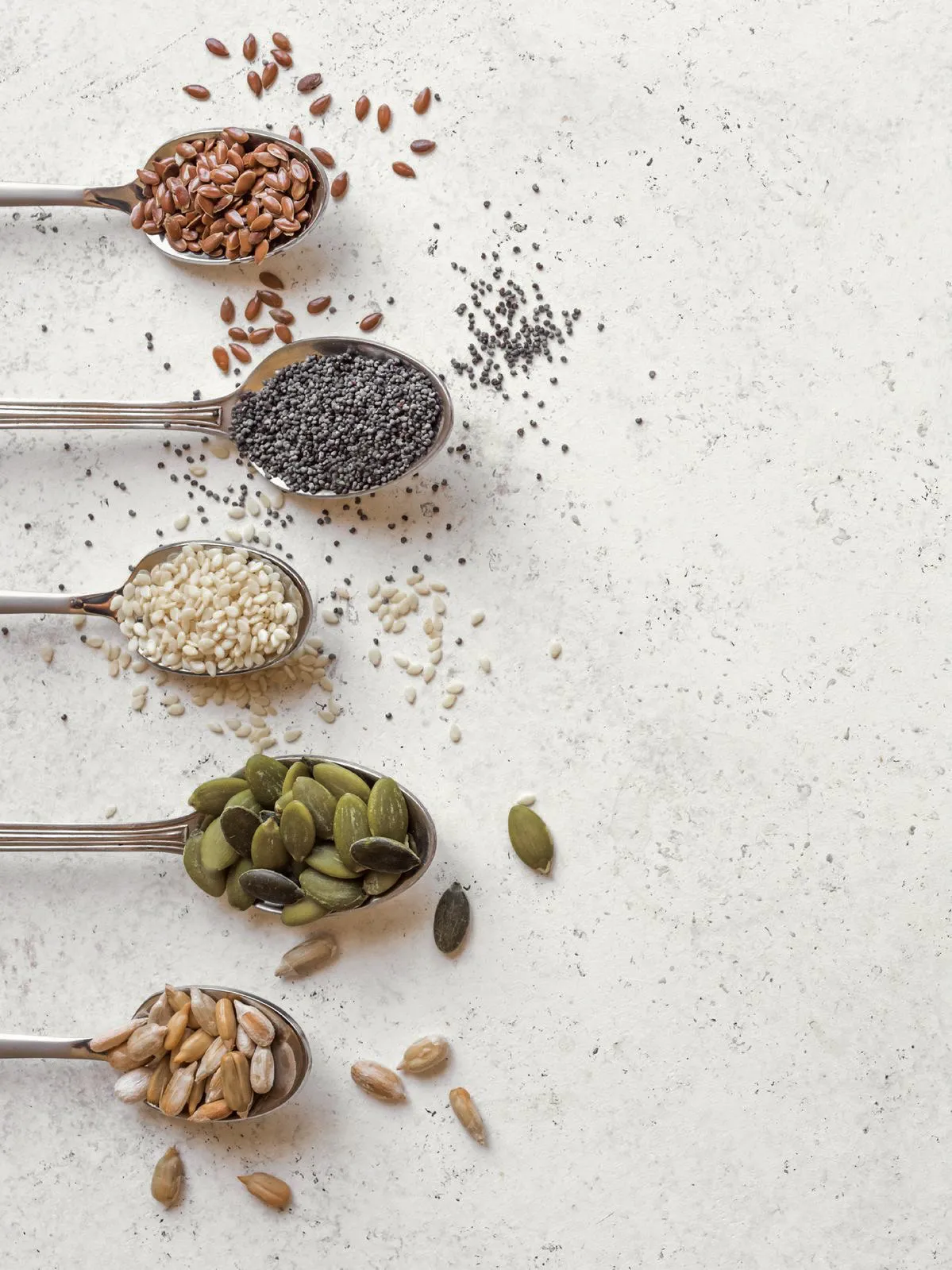 Five spoons of seeds — flax, poppy, sesame, pumpkin, sunflower — on stone.