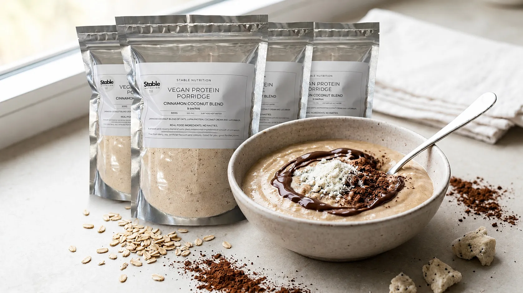 Stable Nutrition Vegan Protein Porridge pouches beside a creamy cinnamon-coconut porridge bowl with a chocolate swirl, oats scattered on the counter.